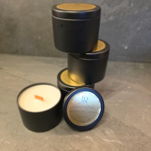 May include: A stack of four matte black metal candle tins with gold circular labels on the lids. One open candle tin sits in the foreground, revealing white wax and a wooden wick. The gold labels feature the text FR velvet trespass ATMOSPHERE CANDLE 4 OZ 113G W.