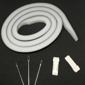 Op de afbeelding: Een witte schuimrubberen weerstrip rol met een zwarte achterkant, vier naalden en twee witte plastic clips.