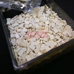 以下が含まれることがあります： 中央に小さな穴が開いた、小さな白いプラスチック製の四角い部品がたくさんあります。部品は透明なプラスチック容器に入っています。