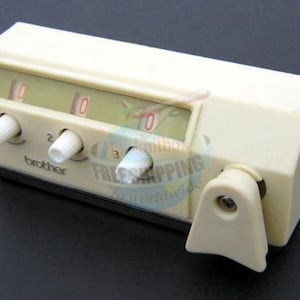 以下が含まれることがあります： 3つのダイヤルとレバーが付いた白いブラザーミシンアクセサリー。ダイヤルには0、1、2、3の数字が書かれています。レバーには「ブラザー」と書かれています。