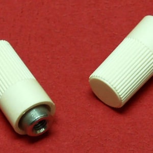 May include: Two off-white cylindrical knobs with ridged sides and a flat end. The knobs have a metal insert on one end. The knobs are set against a red background. These knobs could be used for furniture or other applications.