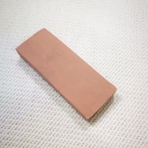 May include: A rectangular, reddish-brown sharpening stone, possibly for knives or tools, lies on a textured white surface. The stone is a long, narrow rectangle with a slightly rough texture. The background is a white, woven fabric.