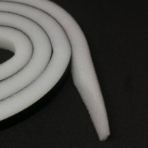 Op de afbeelding: Een close-up van een witte schuimrubberen weerstrip met een zwarte kleefrug. De tape is opgerold en het uiteinde is naar buiten gekruld.
