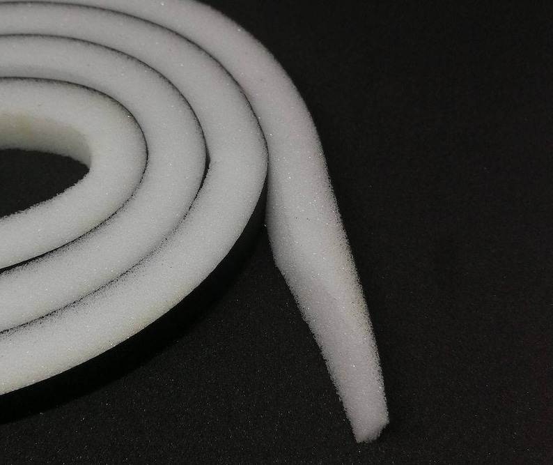 Op de afbeelding: Een close-up van een witte schuimrubberen weerstrip met een zwarte kleefrug. De tape is opgerold en het uiteinde is naar buiten gekruld.