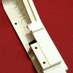 May include: A white plastic part for a sewing machine. The part has a curved shape and several holes. The text "SCKMCF" is visible on the part.