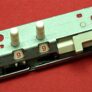 May include: A rectangular metal component with three white knobs and two windows displaying the number "9" in red. The component also has a white sliding mechanism and is set against a red background.