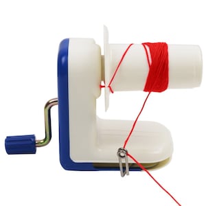 May include: A white and blue yarn winder with a red yarn being wound onto the spool. The winder has a crank handle and a clip to hold it in place.