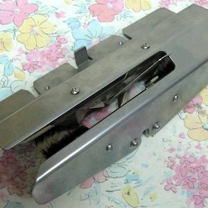 May include: A stainless steel sewing machine attachment with a rectangular body and several protruding tabs. The attachment has a slot revealing internal components and is secured with small screws. The object is designed for sewing.