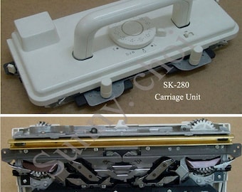 May include: White knitting machine carriage unit with a handle and a dial. The carriage is open to show the internal workings, including gears and metal parts.