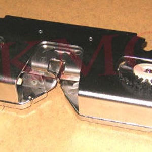 May include: Close-up of a silver-toned metal sewing machine foot with two black brushes. The foot has a small red button on the side.