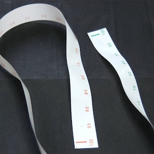 May include: Two white measuring tapes with red and green markings. The tapes are marked in increments of 5, starting at 0 and ending at 100. The tapes are lying on a black surface.