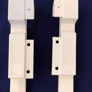 May include: Two white plastic corner brackets with a square shape and a small hole on each side. The brackets are designed to connect two pieces of trim or molding at a 90-degree angle.