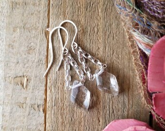 Puede incluir: Pendientes colgantes de plata con colgantes de cristal transparente facetado. Los pendientes presentan un diseño de cadena y se muestran sobre una superficie de madera rústica. Los cristales están tallados en formas geométricas.
