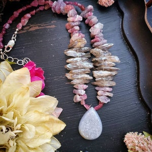 May include: A pink and grey gemstone necklace with a teardrop-shaped pendant. The necklace is made of faceted beads and has a silver clasp.