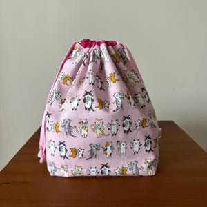 May include: A pink drawstring bag featuring a pattern of cats enjoying ice cream cones. The bag is made from cotton fabric and has a pink drawstring.