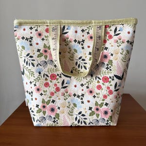 May include: A cream-colored tote bag with a floral pattern featuring pink, red, and blue flowers, and green leaves. The handles are made from a measuring tape. The top edge is trimmed with a light green fabric.