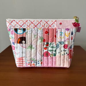 May include: A quilted, rectangular pouch with a zipper closure. The patchwork design features various floral and patterned fabrics in shades of pink, red, and white. A cherry charm adorns the zipper pull. The pouch is approximately 8 inches wide.