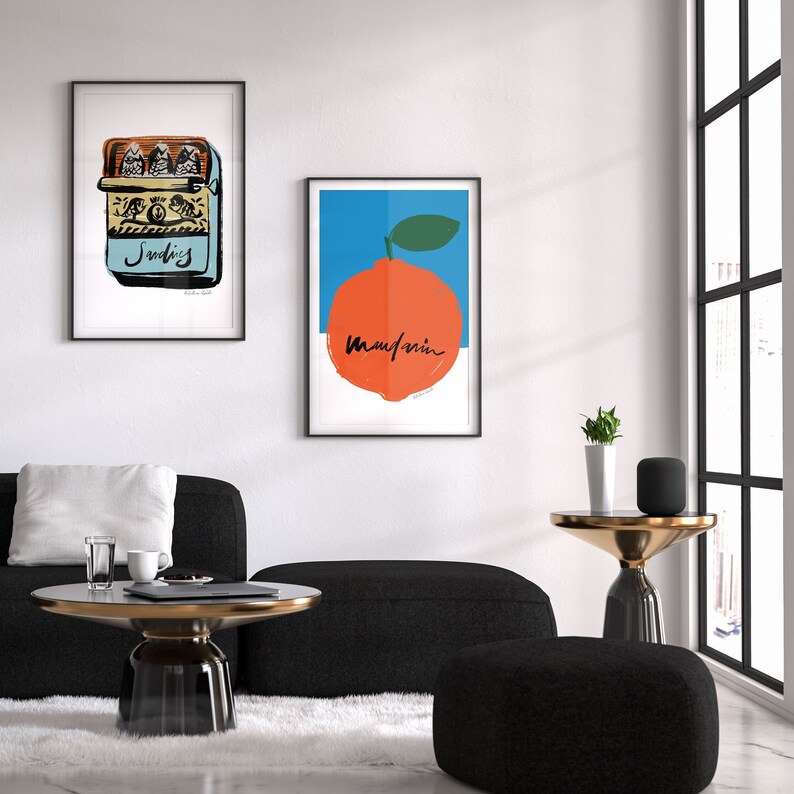 May include: Two framed prints on a white wall. The print on the left is a yellow and blue illustration of a sardine can. The print on the right is a blue and orange illustration of a mandarin orange with the word "Mandarin" written in black.