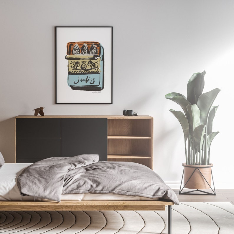 May include: A framed illustration of a vintage sardine can with three sardines inside. The can is labeled "Sardines" and has a blue background with a yellow and orange border. The illustration is in a black frame and is hanging on a white wall.