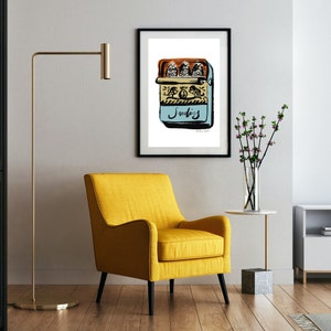 May include: A black framed print of a hand-drawn illustration of a blue can of sardines with three fish inside. The can has the word "Sardines" written on it in black script. The print is hanging on a white wall above a yellow armchair.