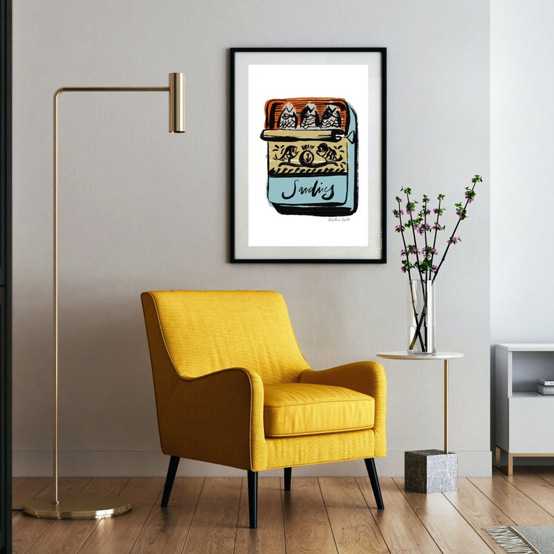 May include: A black framed print of a hand-drawn illustration of a blue can of sardines with three fish inside. The can has the word "Sardines" written on it in black script. The print is hanging on a white wall above a yellow armchair.