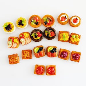 Puede incluir: Una colección de alimentos en miniatura, que incluye tartas, tostadas y pasteles, dispuestos sobre una superficie blanca. Los alimentos están decorados con varias frutas como uvas, fresas y mangos. Los colores son vibrantes y los detalles son intrincados.