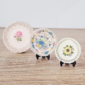 Könnte beinhalten: Drei dekorative Teller mit Blumenmustern. Die Teller sind weiß mit goldenen Verzierungen und zeigen verschiedene Blumenmuster. Die Teller stehen auf kleinen schwarzen Ständern.