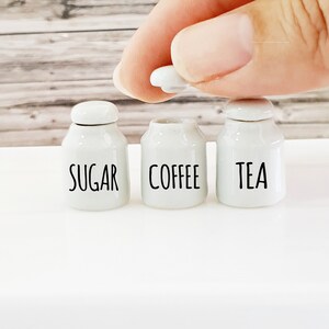 Op de afbeelding: Drie witte keramische potten met zwarte letters. De potten zijn gelabeld met "Suiker", "Koffie" en "Thee".