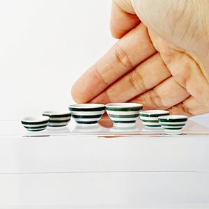 May include: Set of six miniature white bowls with green stripes. The bowls are arranged in order of size, with the largest bowl on the left and the smallest bowl on the right.