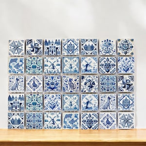 Puede incluir: Una colección de 36 azulejos de Delft azules y blancos con varios diseños, incluyendo molinos de viento, flores, casas y animales. Los azulejos están dispuestos en un patrón de cuadrícula en una pared blanca.