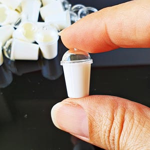May include: Miniature white plastic cup with a clear lid, held between two fingers. Several similar cups are in the background. The cup appears to be filled with a light-colored liquid. The cup is approximately 1 inch tall.