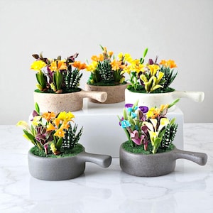May include: Several small, decorative flower arrangements in shades of yellow, orange, purple, and blue. The flowers are planted in small, round, speckled ceramic pots with handles. The pots are in shades of grey and beige.
