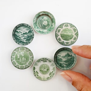 May include: Six small green and white ceramic plates with different floral and landscape designs. The plates are arranged in a circle on a white background.
