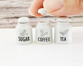 May include: Three white ceramic canisters with black lettering and illustrations. The canisters are labeled "Sugar", "Coffee", and "Tea".