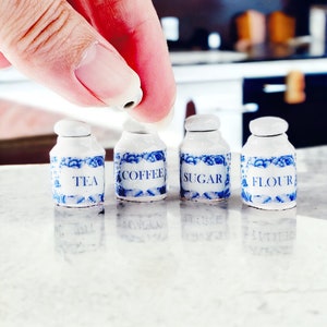 Puede incluir: Cuatro botes de cerámica blanca con diseños florales azules y etiquetas que dicen "Té", "Café", "Azúcar" y "Harina".