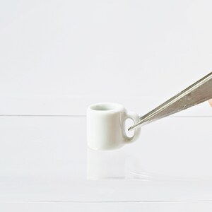 May include: A small white ceramic mug with a handle, held by silver tweezers.