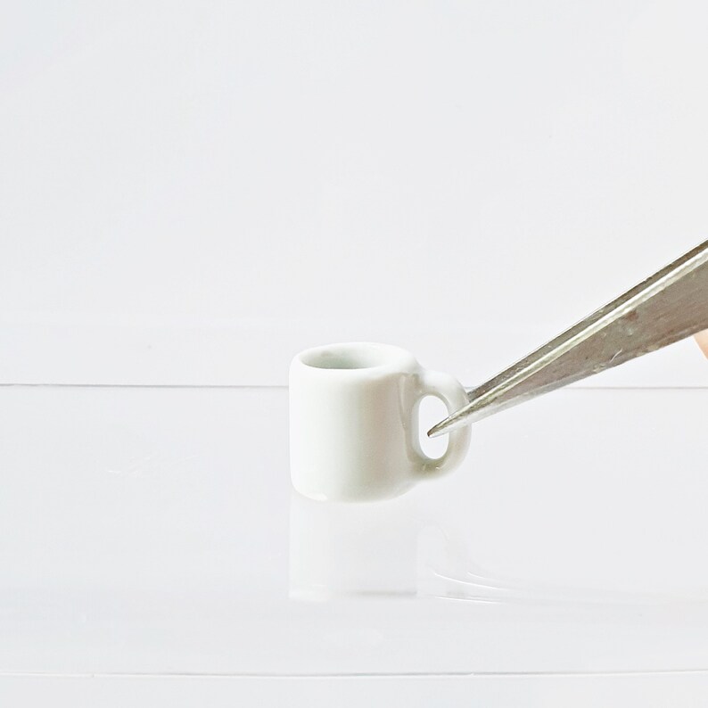 May include: A small white ceramic mug with a handle, held by silver tweezers.