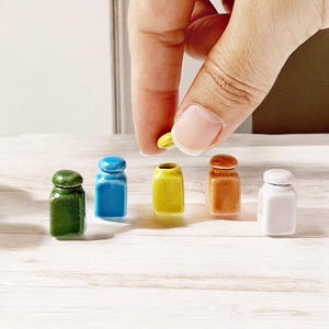 May include: Five miniature ceramic jars in different colors: green, blue, yellow, brown, and white. The jars are lined up on a white wooden surface. A hand is holding a yellow jar.