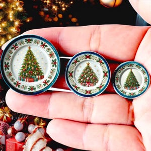 Op de afbeelding: Drie miniatuur decoratieve borden met kerstboomontwerpen. Elk bord heeft een versierde boom met ornamenten en cadeaus, tegen een witte achtergrond met een hulst- en lintrand. De borden hebben een blauwe rand.