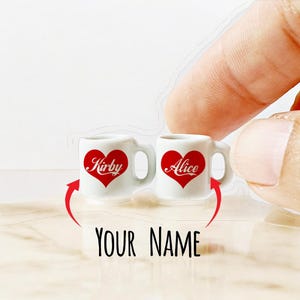 Peut inclure: Deux tasses en céramique blanche avec des cœurs rouges et les noms "Kirby" et "Alice" imprimés dessus. Les tasses sont posées sur une surface blanche avec le texte "YOUR NAME" imprimé en dessous.