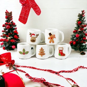 May include: Six white ceramic mugs with Christmas-themed designs, including a reindeer, gingerbread man, holly, Christmas tree, and stocking. The mugs are arranged on a white surface with festive decorations.