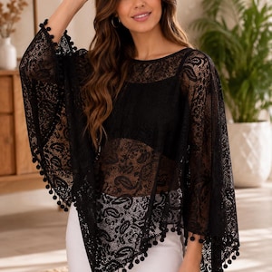 May include: A black lace poncho featuring an intricate paisley pattern and a wide, loose-fitting silhouette. The garment has a round neckline and is finished with small, round black pom-pom trim along the edges of the sleeves and hemline.