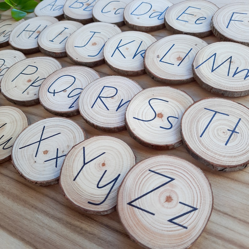 May include: Wooden alphabet letter tiles with black lettering. The letters are arranged in alphabetical order on a wooden surface.