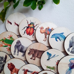 May include: A set of 16 wooden circle game pieces featuring various animal and insect illustrations. The pieces include a camel, a hippopotamus, a dragonfly, a gorilla, a frog, a beetle, a jellyfish, an octopus, a lion, a monkey, a squirrel, a dolphin, an ant, and a few other animals.