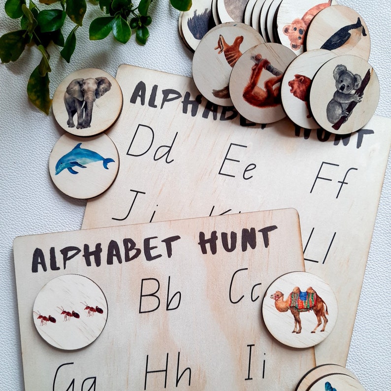 May include: Alphabet Hunt learning game with wooden circles featuring animal illustrations. The circles are placed on two wooden boards with the letters of the alphabet written on them. The boards say "Alphabet Hunt".