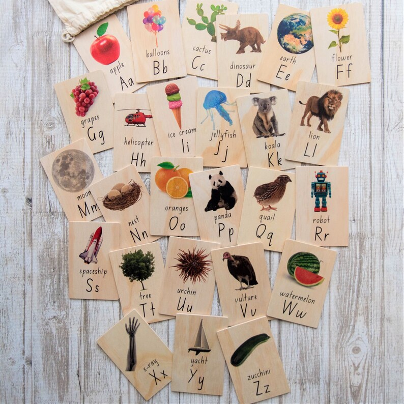 May include: A set of wooden flashcards, each displaying a letter of the alphabet alongside a corresponding image. The illustrations include an apple, balloons, cactus, dinosaur, earth, flower, grapes, helicopter, ice cream, jellyfish, koala, lion, moon, nest, oranges, panda, quail, robot, spaceship, tree, urchin, vulture, watermelon, x-ray, yacht, and zucchini.