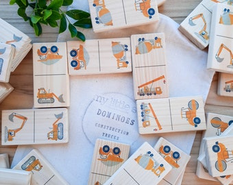 May include: A set of wooden dominoes featuring illustrations of construction vehicles. Each domino has two halves, with a different vehicle on each half. The vehicles are all yellow and orange and include a cement mixer, a crane, a bulldozer, and an excavator. The dominoes are labelled "My Little Dominoes Construction Trucks".