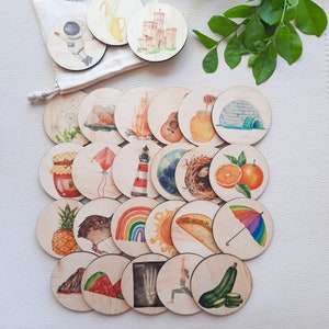 Puede incluir: Un conjunto de 20 fichas circulares de madera con ilustraciones de acuarela. Las fichas presentan una variedad de im&aacute;genes, incluyendo una pi&ntilde;a, un faro, un arco&iacute;ris, un nido con huevos, una fogata, una guitarra, una rodaja de sand&iacute;a y un igl&uacute;.