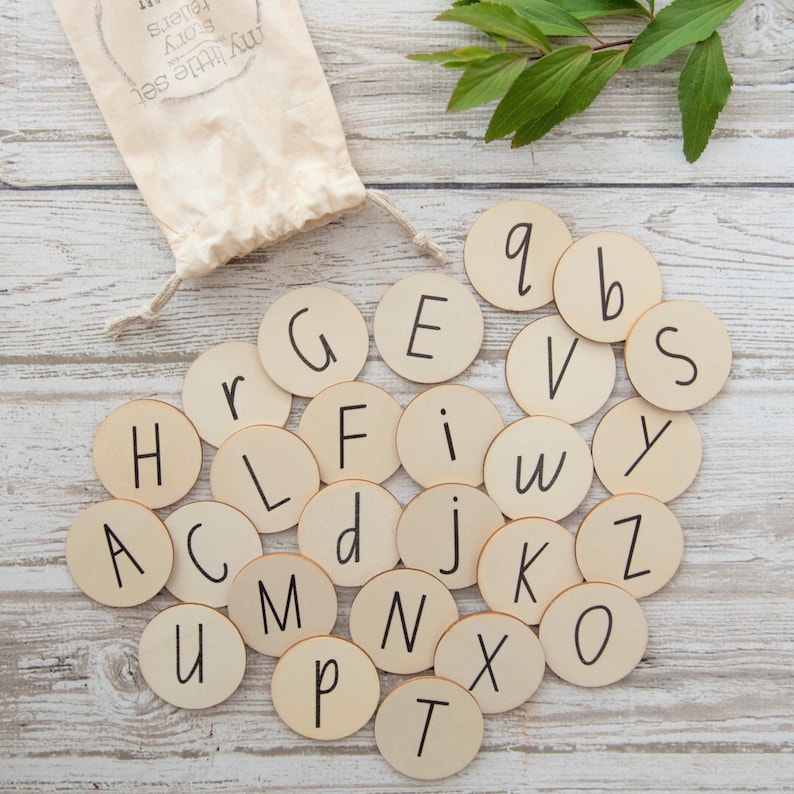 May include: A set of wooden alphabet letter tiles in a white drawstring bag. The letters are painted black on a light wood background. The letters are arranged in a circle with the bag in the centre.