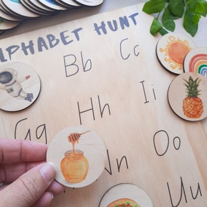 Puede incluir: Juego de mesa de madera Alphabet Hunt con fichas de letras que presentan im&aacute;genes de un tarro de miel, una pi&ntilde;a, un sol, un arco&iacute;ris y un astronauta. El tablero tiene letras escritas en &eacute;l, incluyendo Bb, Cc, Hh, Ii, Oo y Uu.